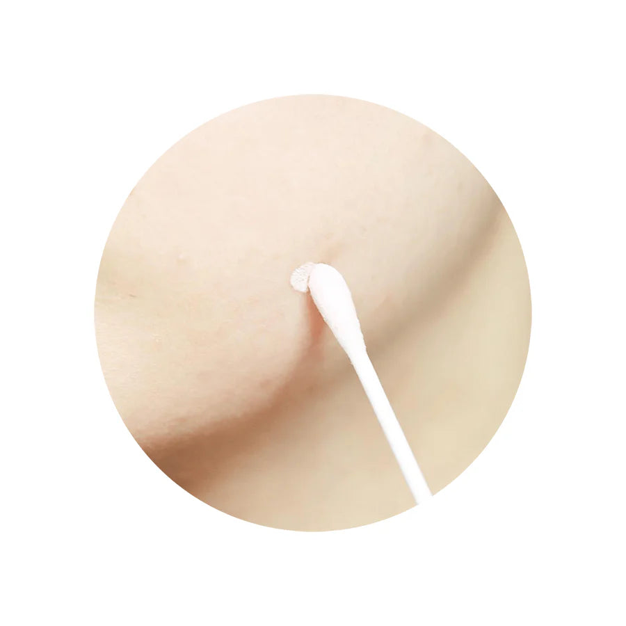 A cotton swab dipping into the pink sediment at the bottom of the bottle, applying the soothing treatment to a targeted acne spot on the skin.